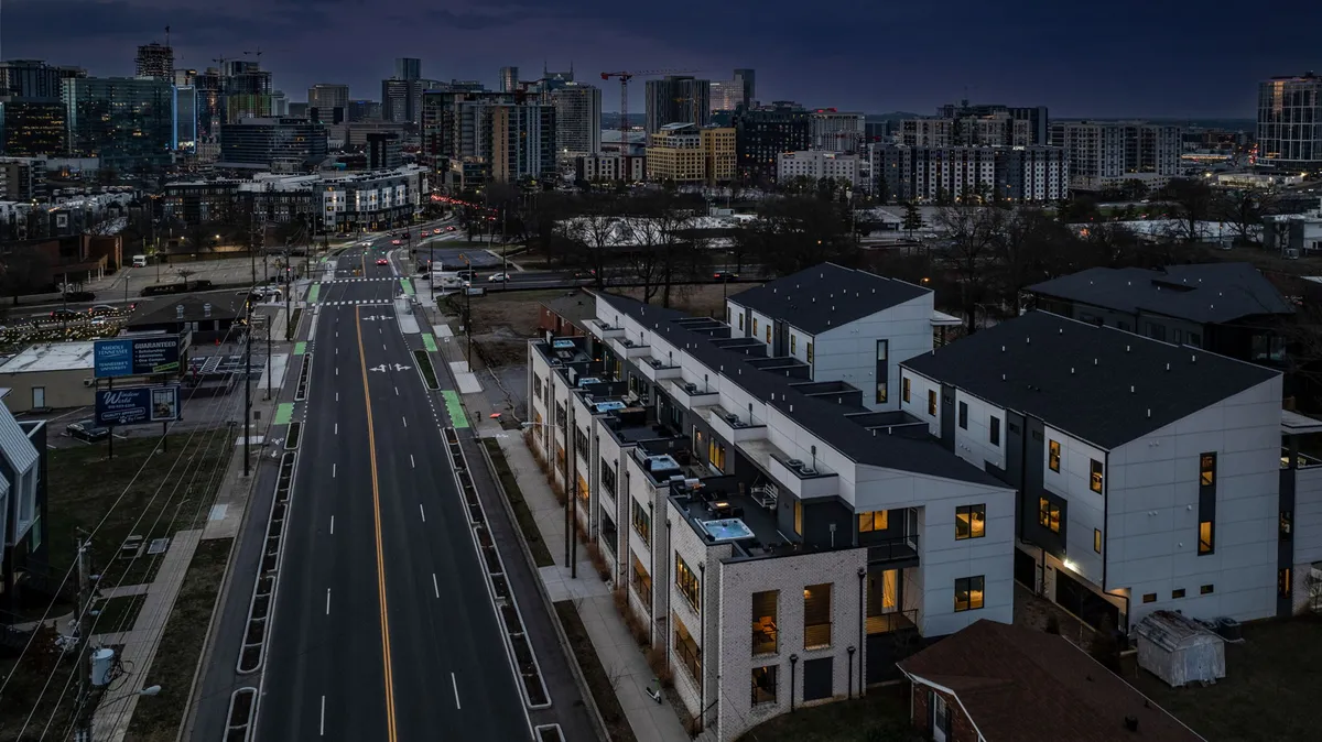 A contemporary cityscape with a row of modern townhouses, a city skyline in the distance, and a walkable street with bike lanes. The atmosphere is vibrant and urban yet welcoming, hinting at the lively neighborhood environment.