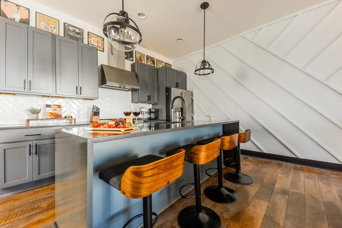 A warm and welcoming modern kitchen featuring a prominent kitchen island with wooden bar stools, sleek cabinets, artwork on the walls, and ample lighting. The space is designed for both cooking and entertaining, with plenty of countertop space and a contemporary ambiance that invites group gatherings.