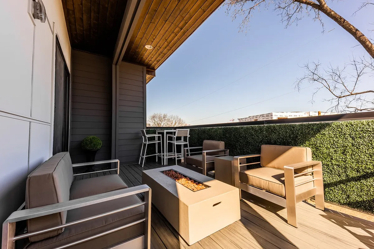 The image features a stylish rooftop deck with modern outdoor seating arranged around a fire pit. Lush greenery softens the space, and a bar-height table offers a spot for drinks or casual dining. The view suggests an elevated vantage point, ideal for sunset watching or stargazing, and the setting is intimate yet open, inviting both small gatherings and quiet moments.