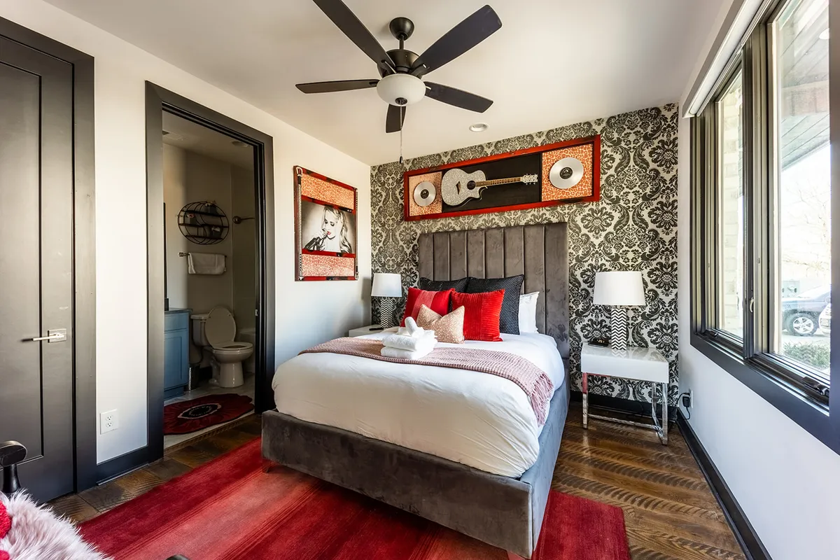 A welcoming bedroom with plush bedding, decorative pillows, guitar-themed wall art, and stylish bedside lamps. The artistic decor references Nashville's musical heritage while providing a cozy, restful atmosphere.