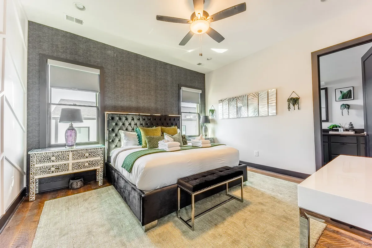 A stylish bedroom featuring a plush bed with decorative pillows, a sleek nightstand, large mirrors, and artistic wall decor. The room is bathed in soft natural light, creating a calm and sophisticated ambiance that feels both inviting and private.
