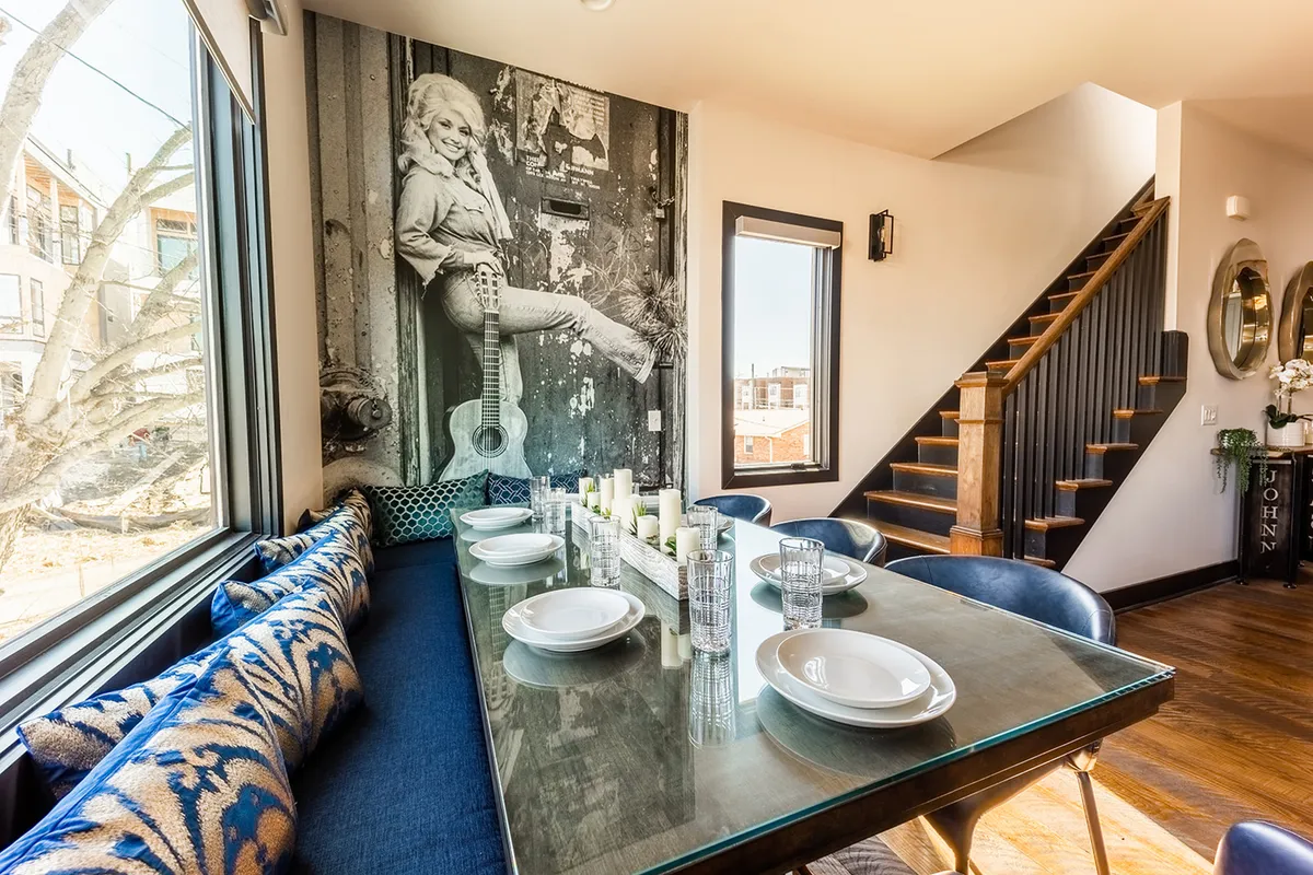 A modern dining area with a glass dining table set with blue patterned cushions and a striking wall mural of a woman with a guitar, evoking a sense of local artistic flair. The space is bright and inviting, perfect for group dining or celebration.
