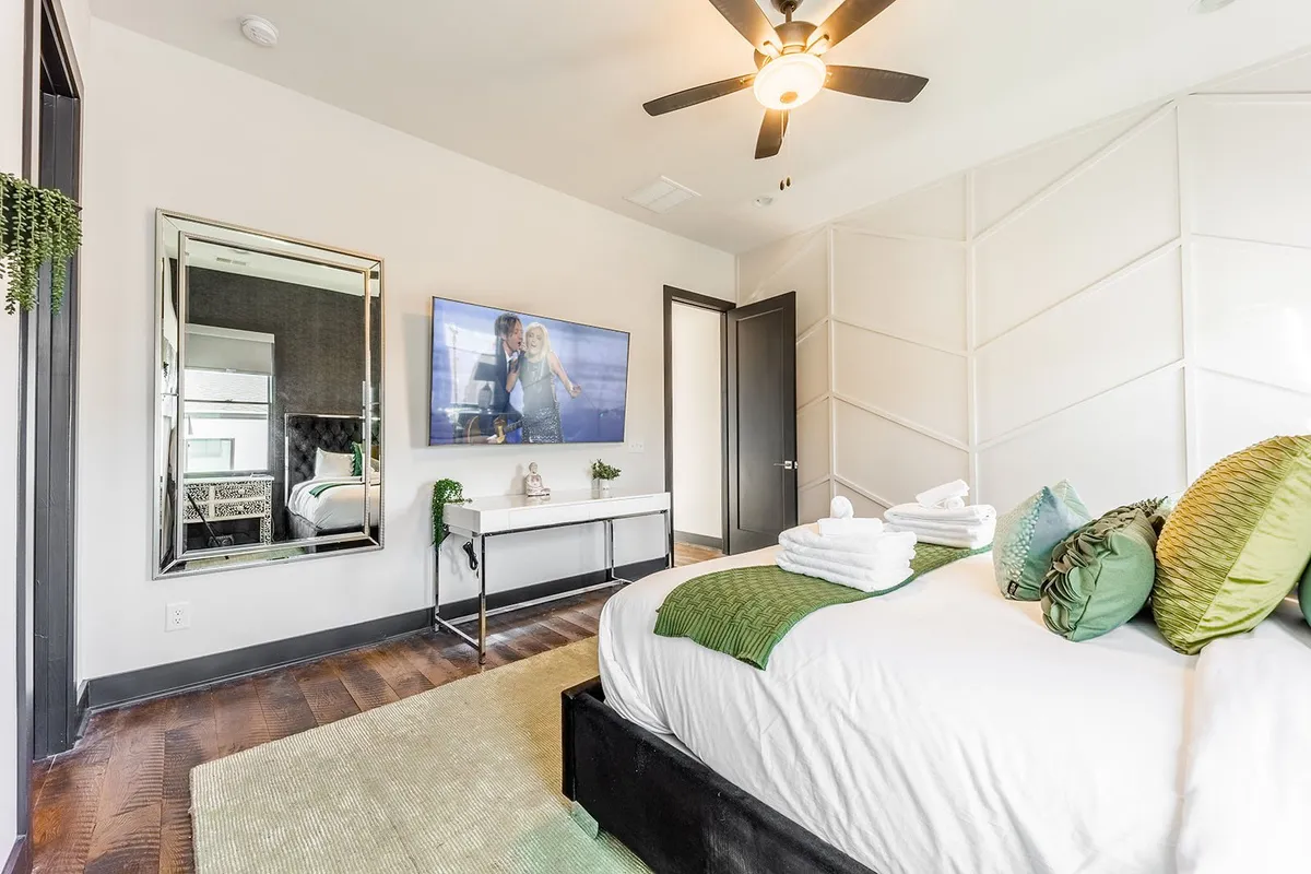 A modern bedroom with a cozy bed adorned with decorative pillows, a wall-mounted television displaying a couple, and a mirror reflecting the room's tranquil atmosphere. The scene suggests relaxation and personal leisure, emphasizing entertainment and comfort.