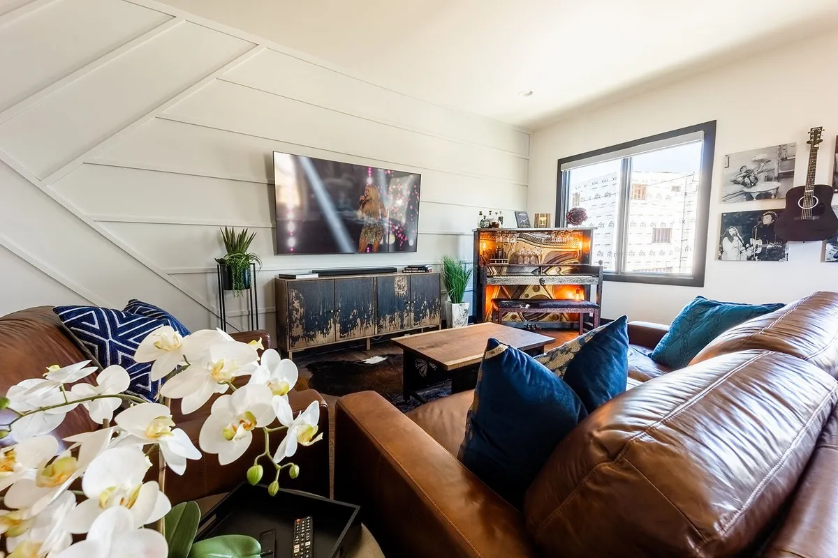 A cozy living room featuring leather sofas arranged around a coffee table, a large flat-screen TV mounted on the wall, a decorative shelf, and a window offering natural light. The space is accessorized for both comfort and style, ideal for gatherings or relaxation.