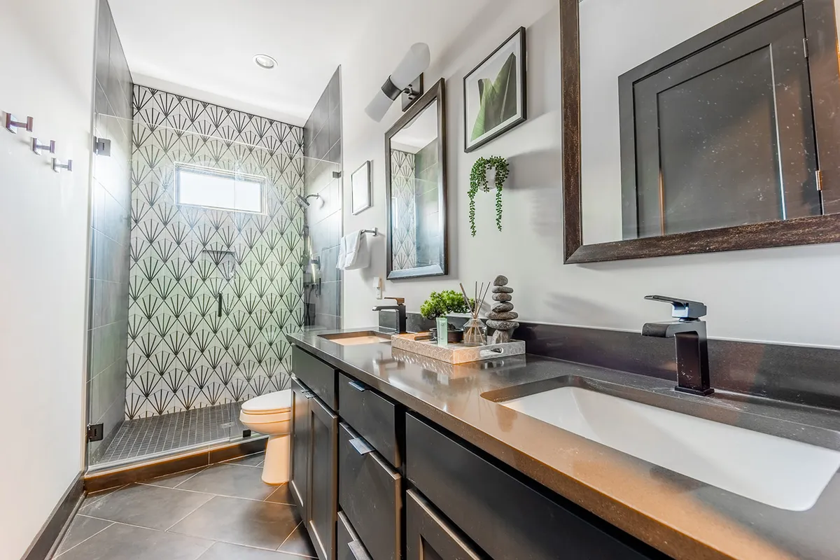 A spa-inspired en-suite bathroom featuring a double vanity with dual sinks, framed mirrors, a decorative geometric tile wall, and a spacious shower area. The space feels calm, luxurious, and meticulously clean.