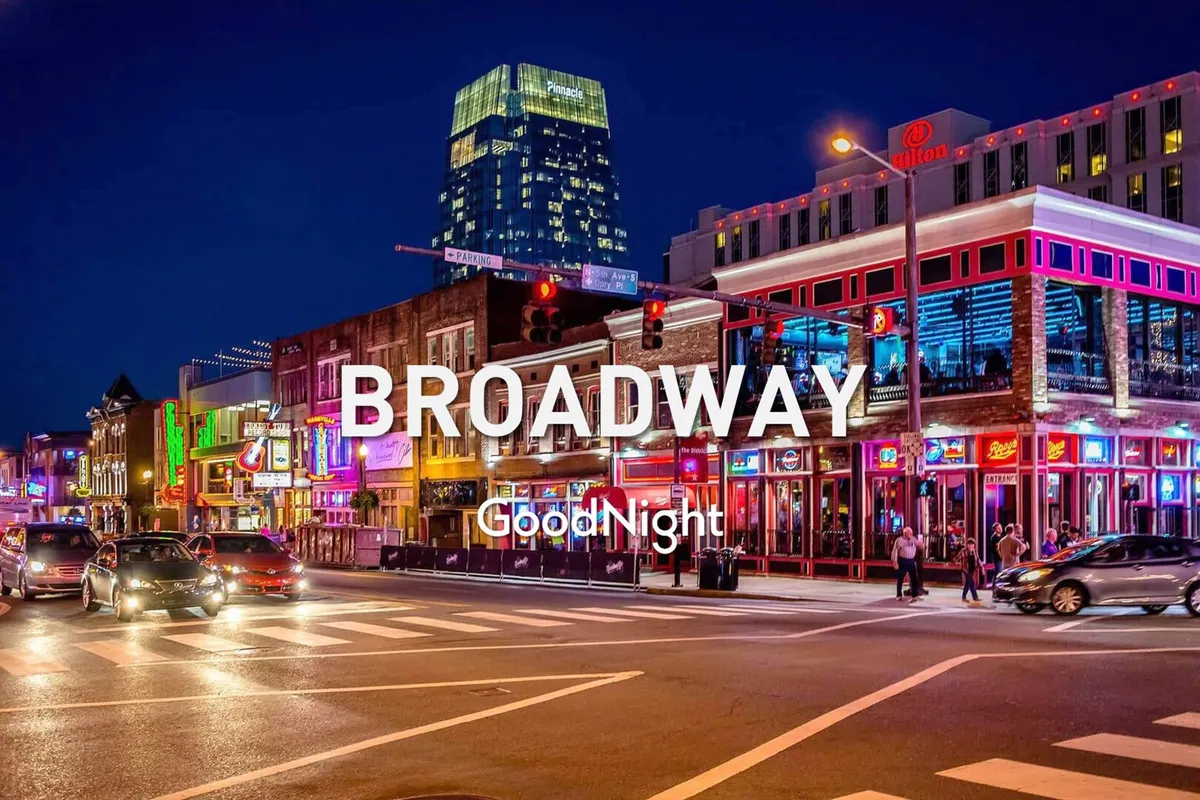 A lively exterior city scene at night, featuring neon signs, a busy crosswalk, high-rise buildings, cars, and pedestrians. The vibrant lights and crowds evoke the legendary energy of downtown Nashville and Broadway's nightlife.