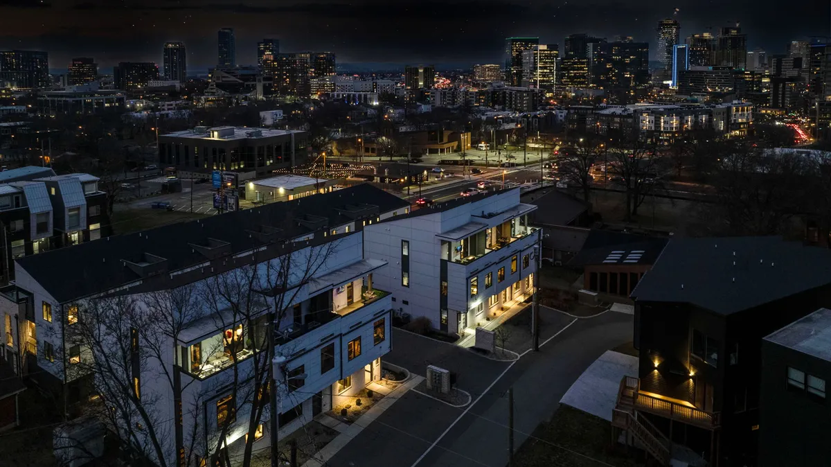 This image features modern townhouses with the Nashville city skyline in the background and bright street lights that signal a lively evening atmosphere. The sidewalks and illuminated facades evoke urban energy, suggesting walkability and proximity to nightlife and dining.