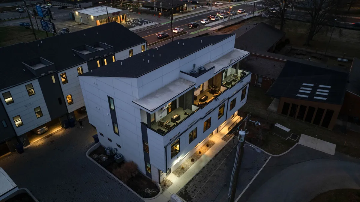 A contemporary exterior shot highlighting the upper floor outdoor seating area, softly illuminated in the evening. The modern architecture and lighting create a warm atmosphere, suggesting a welcoming environment for social gatherings, alfresco dining, and enjoying the city skyline at dusk.