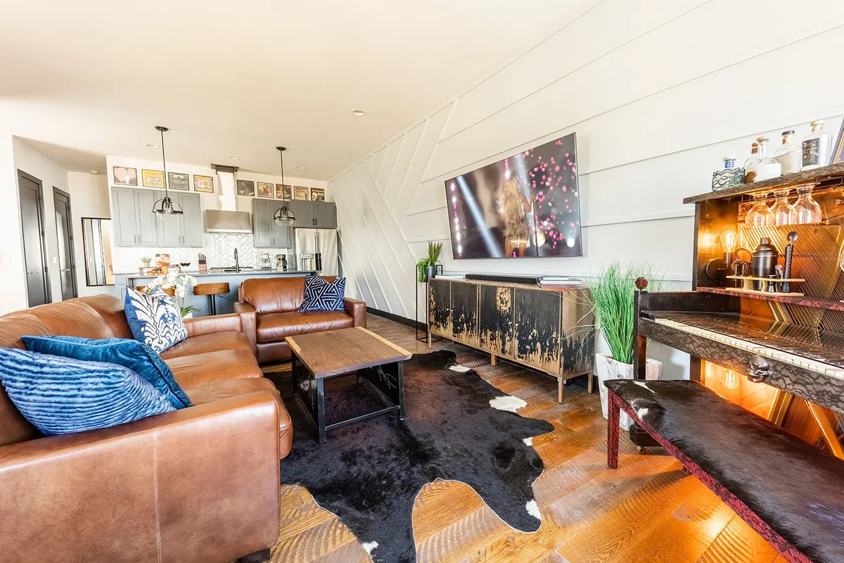 The image showcases a chic, open-concept living room with a plush leather sofa, sleek coffee table, large television, and a striking bar area—perfectly designed for entertaining and relaxation. The space exudes warmth and sophistication, inviting guests to gather, unwind, and celebrate in comfort.